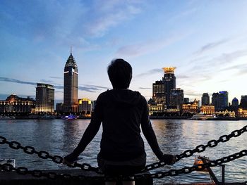 Rear view of woman sitting by river against cityscape