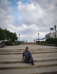 Man sitting on sidewalk in city against sky