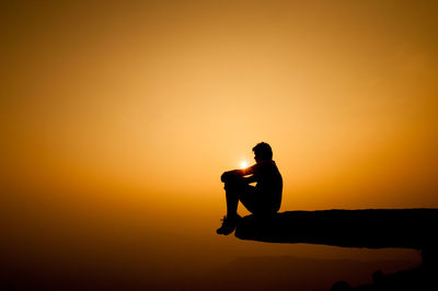 Silhouette man sitting on cliff against sky during sunset