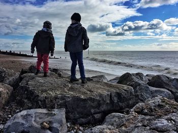 Rear view of brothers standing on rock at beach against cloudy sky during sunny day