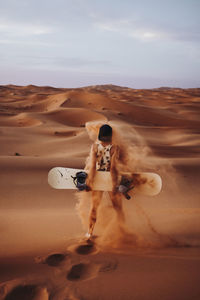 Back view of unrecognizable young female standing on sand with sand over her and preparing for sandboarding