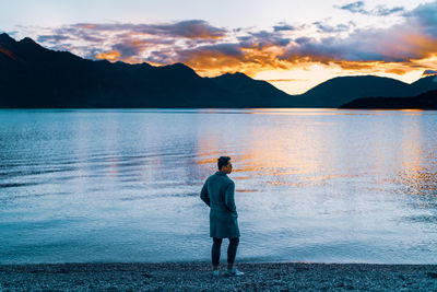 Rear view of man standing on lake against sky during sunset