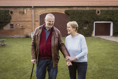 Smiling senior woman holding hand of man walking with cane at lawn in back yard