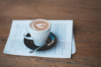 Cute cup of coffee with heart on milk froth staying on open magazine on wooden tale in kitchen close