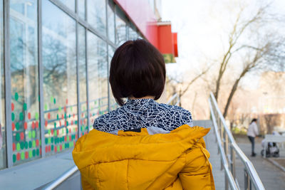 A brunette woman walks through the city with bright orange jacket shoulders, a view from the back.