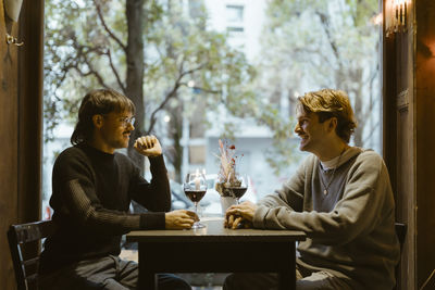 Happy gay couple talking while sitting with wineglasses on table during date at restaurant