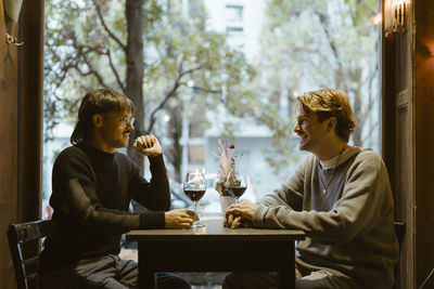 Happy gay couple talking while sitting with wineglasses on table during date at restaurant