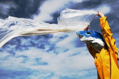 Low angle view of person wearing mask holding textile against sky