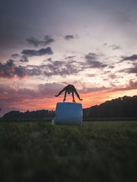 Silhouette man jumping over hay bale against sky during sunset