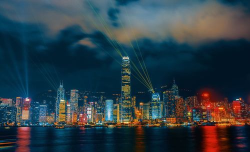 Illuminated buildings by sea against sky at night