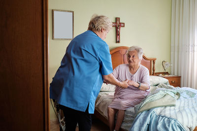 Anonymous nurse in uniform helping elderly female in nightwear to get out of bed with blanket in light bedroom at home