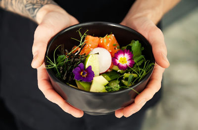 A man holds a bowl of poke salad with salmon, avocado, cucumber, rice, tamarind sauce, mango sauce