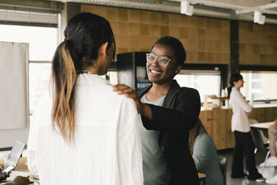 Happy businesswoman with hand on non-binary colleague's shoulder at office