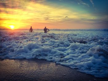 Surfers with surfboard walking on beach against sky during sunset