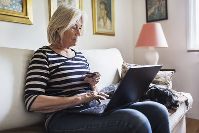 Senior woman shopping online while dog relaxing on sofa at home