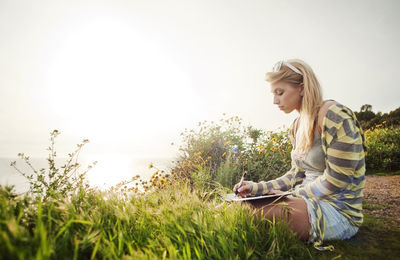 Side view of woman writing while sitting on field against clear sky