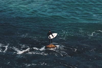 Rear view of man with surfboard standing on rock amidst sea