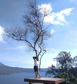 A woman was standing near a tree looking at the view of the lake and the blue sky