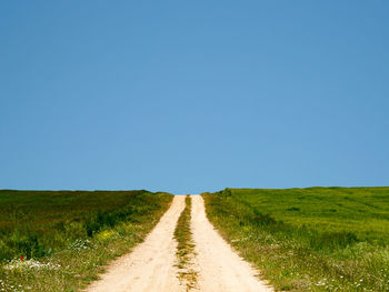 Dirt road amidst field against clear blue sky