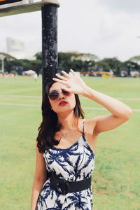 Young woman wearing sunglasses shielding eyes while standing on grassy field in park