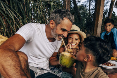 Smiling mature man drinking coconut water with son and daughter on vacation