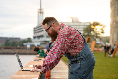 Brutal bearded man freelancer working on laptop at embankment outdoors. distance job, remote work.