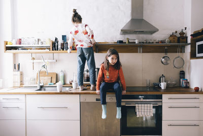 Full length of girl sitting by sister searching on shelf while standing on kitchen counter at home