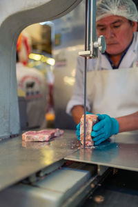 Mature man in apron using electric tabletop saw to slice piece of frozen meat during work in butchery