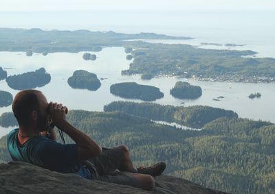 Man looking at lake through binoculars while relaxing on cliff