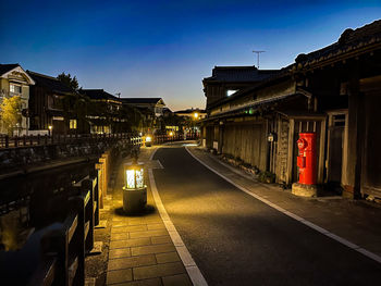 Illuminated street amidst buildings against sky at dusk