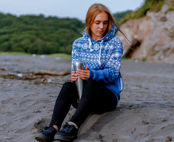 Young woman holding insulated drink container while sitting at beach