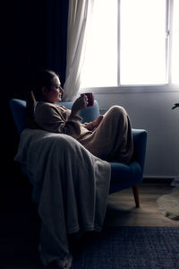 Young woman sitting on sofa at home and drinking coffee or tea