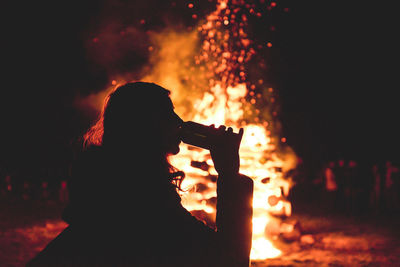 Silhouette woman drinking drink while standing against bonfire