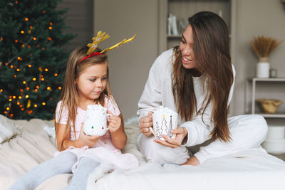 Cute funny little girl with mother on bed in room with christmas tree at home 