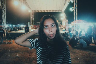 Portrait of beautiful young woman sticking out tongue against stage at night