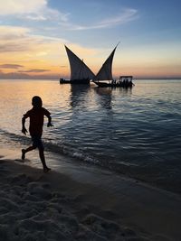 Full length of silhouette boy running at beach by sailboats against sky during sunset