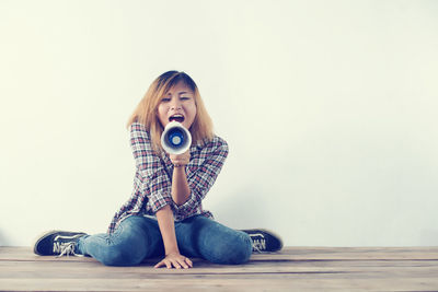 Young woman shouting on megaphone while sitting against white wall