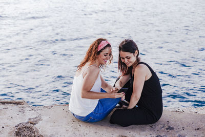 High angle view of lesbian couple using phone while sitting by sea