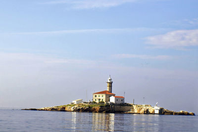 Lighthouse amidst buildings by sea against sky