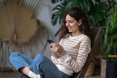 Smiling woman sit in armchair in indoor garden relaxed chatting in social media using smartphone