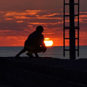 Silhouette person crouching on retaining wall against red sky during sunset