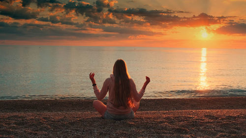 Rear view of woman sitting on beach against sky during sunset