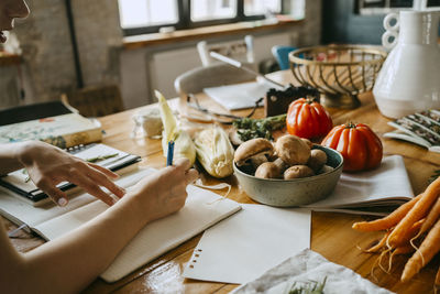 Hand of female food stylist writing in diary by fresh vegetables on table in studio