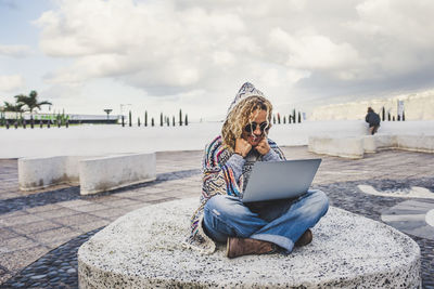 Full length of woman sitting on mobile phone against sky