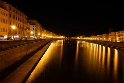 Illuminated street by river and buildings against sky at night