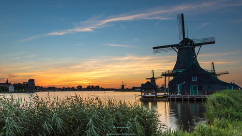 Traditional windmill by lake against sky during sunset