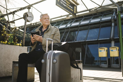 Businessman looking away holding smart phone while sitting on bench by luggage at railroad station