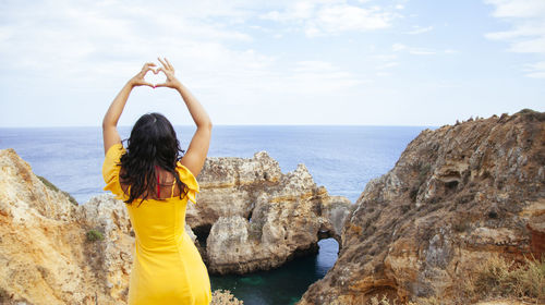 Rear view of woman making heart shape standing by rock against sky