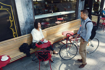 Businessman with bicycle talking to female colleague sitting at sidewalk cafe