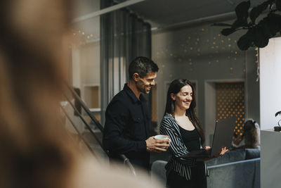 Happy businesswoman sharing laptop with male coworker holding coffee cup at workplace